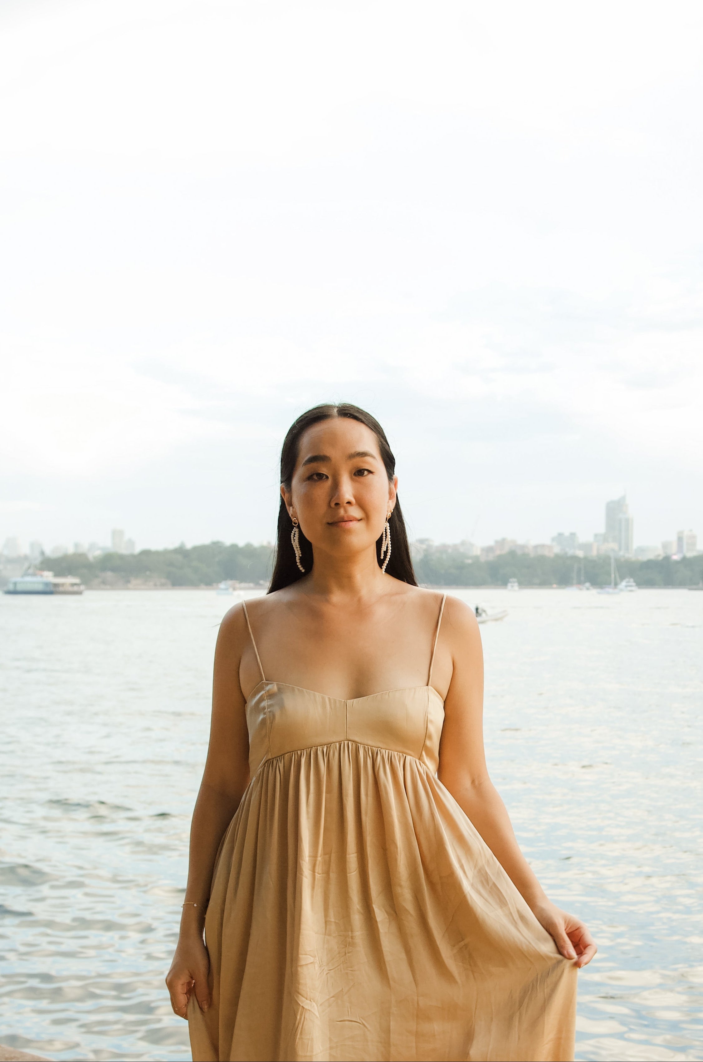 Woman in a long beige dress standing by a waterfront with a city skyline in the background