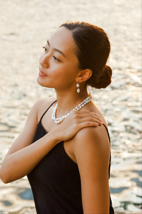 Woman in a black dress sitting by a body of water, with double drop pearl earrings and double tier pearl necklace.