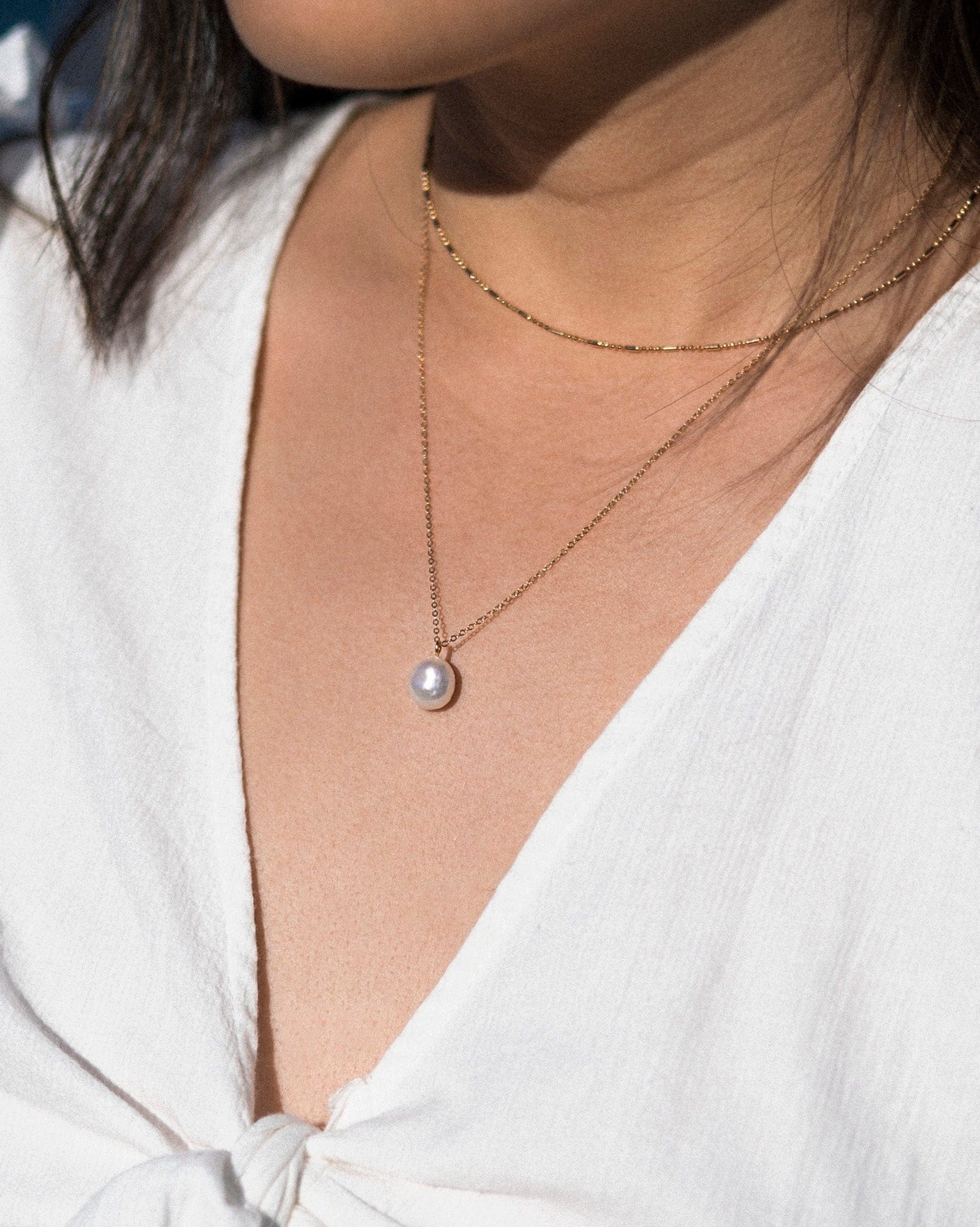 A person wearing a white top with a V-neckline, showcasing a gold-colored necklace with a pearl pendant.