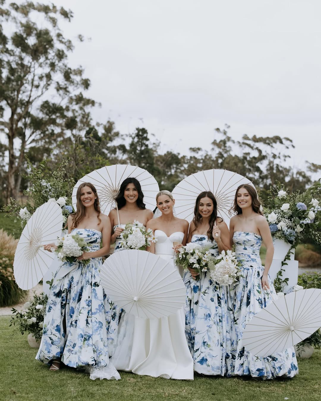 Group of women in floral dresses with a central bride holding a bouquet, surrounded by decorative parasols and flowers outdoors.
