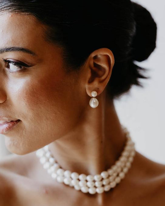 A woman in profile wearing a pearl strand necklace and matching pearl stud earrings, with her dark hair styled in a low bun.